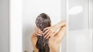 Woman rinsing long hair under a stream of water in a modern white-tiled shower.