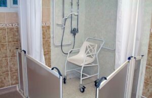 Accessible walk-in shower with safety grab rails, a shower chair on wheels, and half-height doors designed for elderly care and retirement villages.