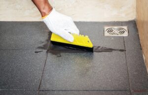 Person using a yellow squeegee to wipe water off a shower floor with visible grout lines and a drain nearby.