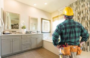 Tradesperson in a yellow hard hat standing in a modern bathroom, looking at the fixtures and renovation details